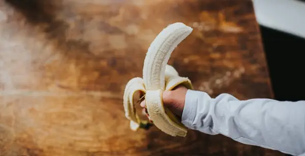Hand holds a peeled banana over a rustic wooden table. 