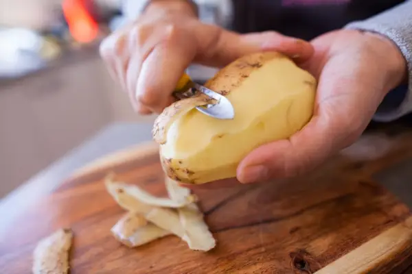 Hands peeling a large potato over a wooden cutting board.