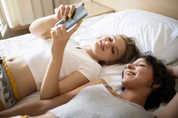 Two women lie on a bed smiling and looking at a mobile phone screen.